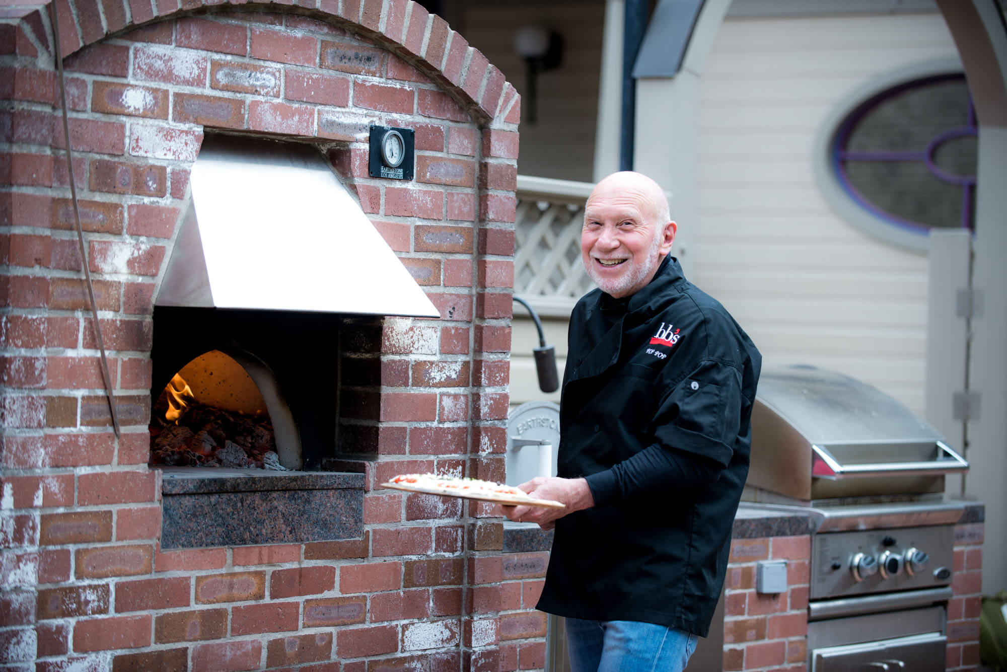 headshot of an older man cooking pizza in an outdoor oven